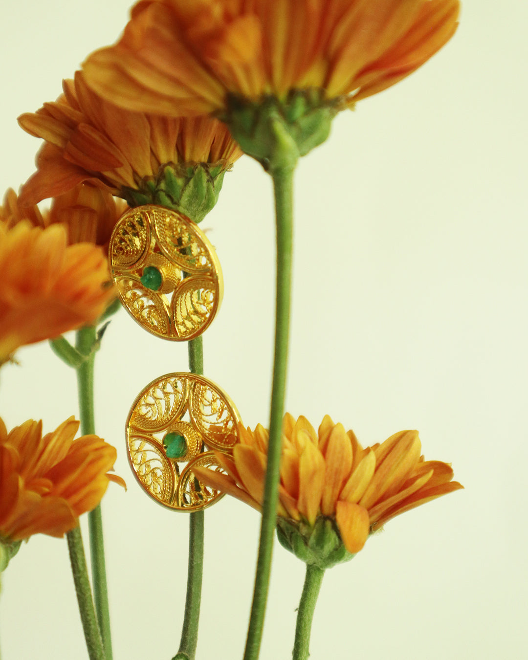 Con Cariño Earrings - Round gold filigree stud earrings with green center stones displayed among vibrant orange chrysanthemum flowers, showcasing intricate handmade Colombian craftsmanship.