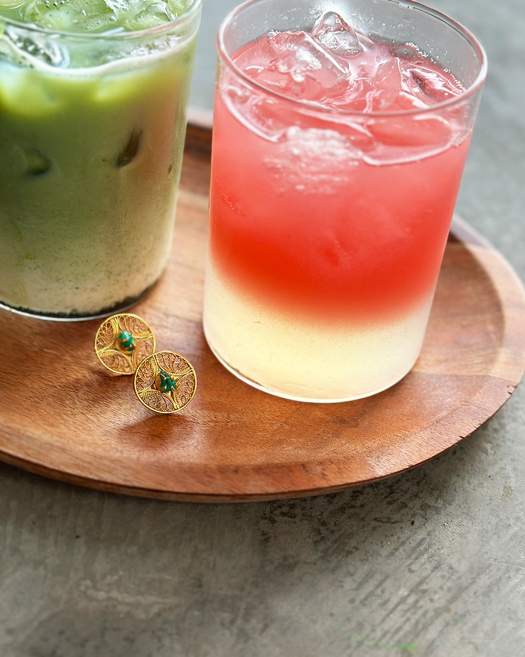 Con Cariño earrings - Round gold filigree earrings with green center stones placed on a wooden tray beside iced matcha and a layered pink citrus drink, combining artisanal jewelry with a fresh, modern lifestyle aesthetic.