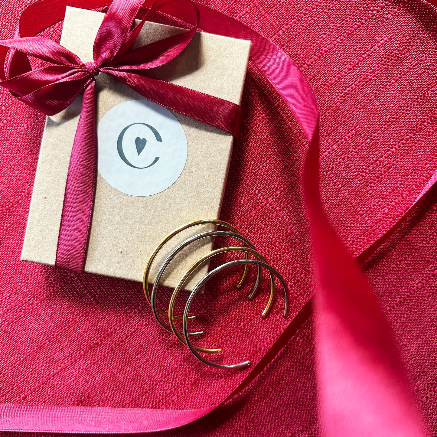 Con Cariño Bracelets - Set of sleek, minimalist gold bangles placed next to a kraft gift box tied with a burgundy ribbon and sealed with a Con Cariño logo sticker, styled on a rich red textured fabric for a festive presentation.