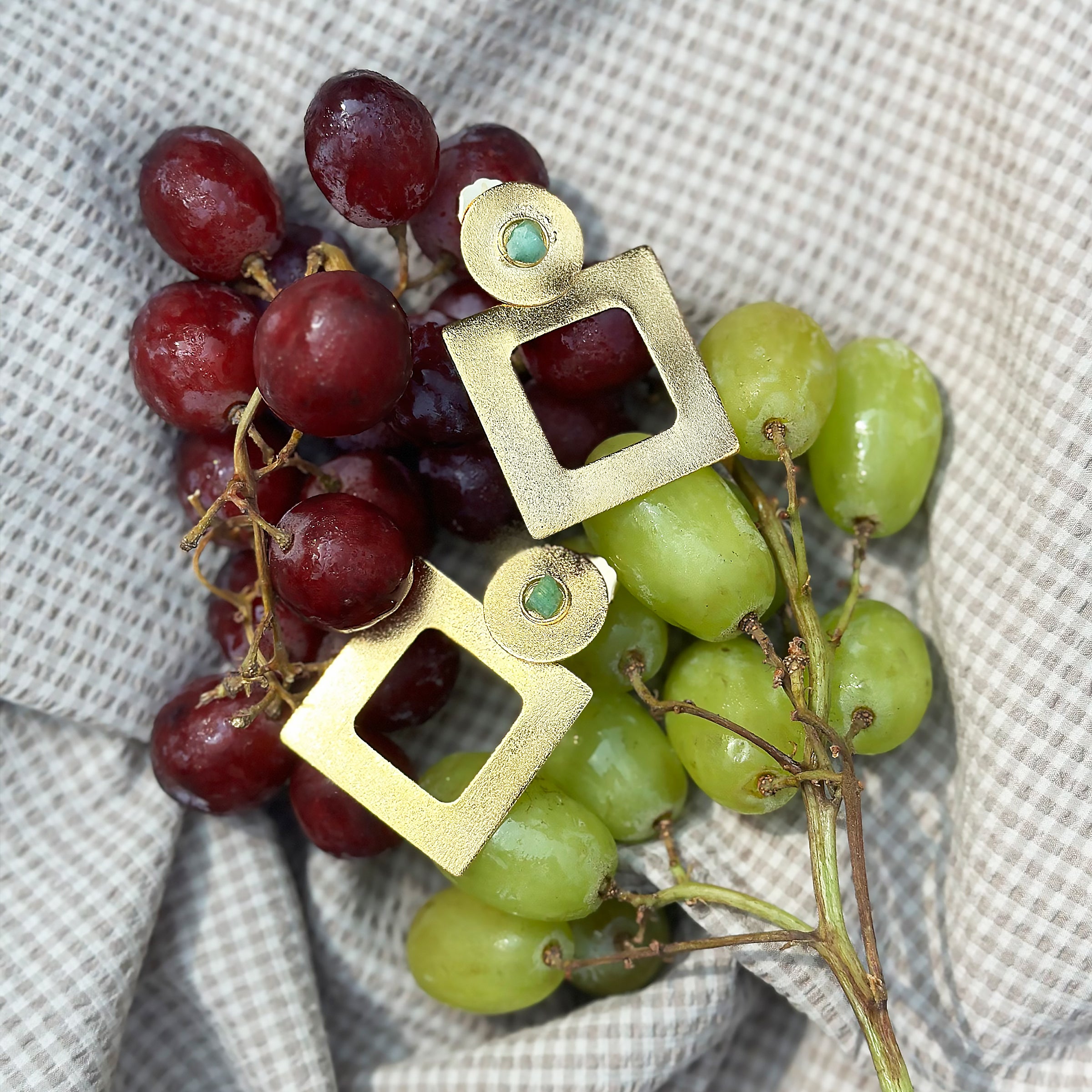A striking pair of Con Cariño Earrings a geometric gold earrings with square cut-out dangles and circular tops, each featuring a small raw emerald, are elegantly placed over a bundle of fresh grapes. The grapes, both red and green, glisten with natural moisture and rest on a soft, checkered beige fabric. The contrast between the vibrant fruit and the matte texture of the gold earrings creates a visually rich and luxurious composition, blending organic beauty with modern artisanal design.