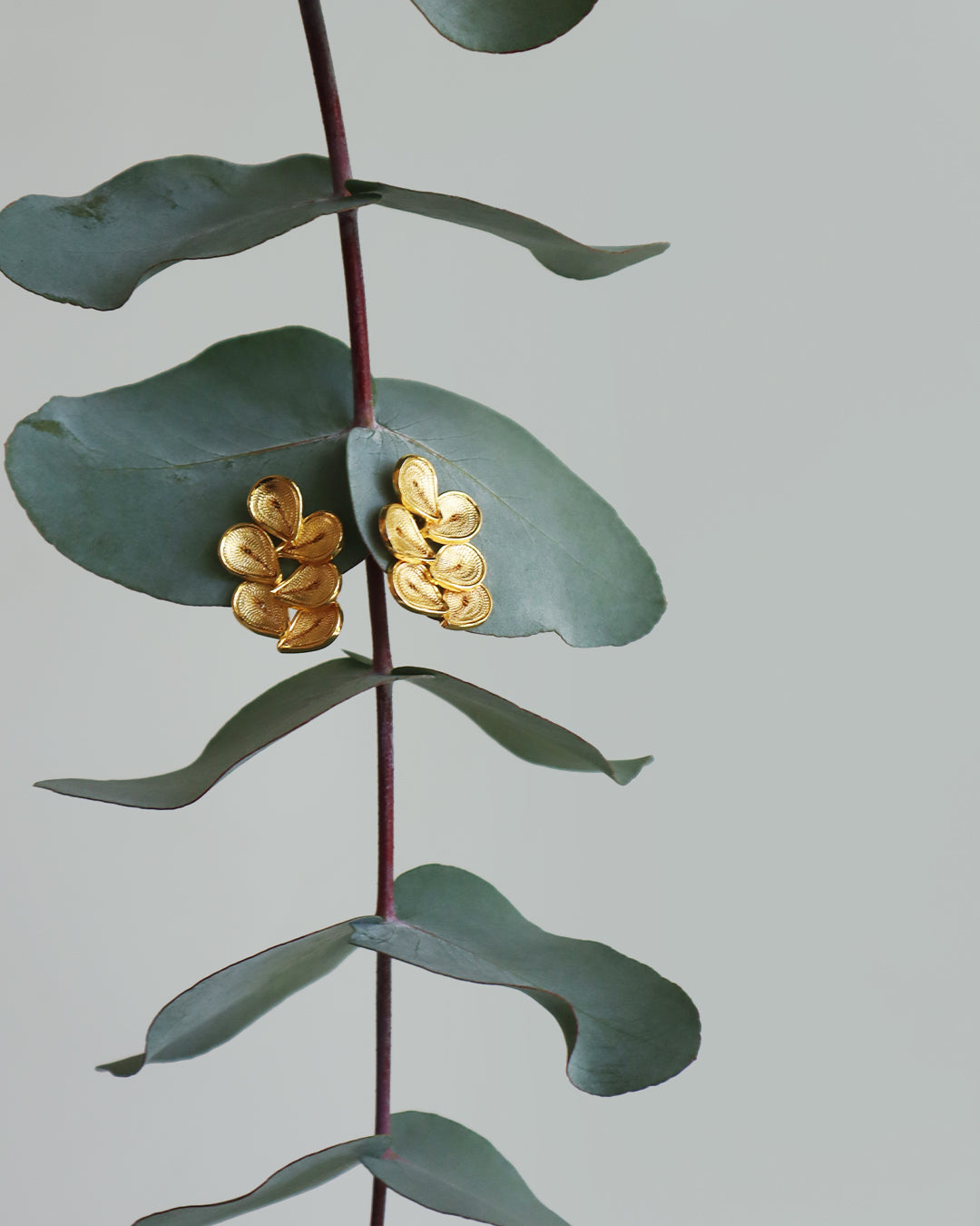 Elegant gold earrings featuring five overlapping teardrop-shaped petals with fine engraved texture, displayed on muted green eucalyptus leaves against a soft gray background, creating a refined and nature-inspired presentation that highlights the handcrafted details and warm metallic finish.