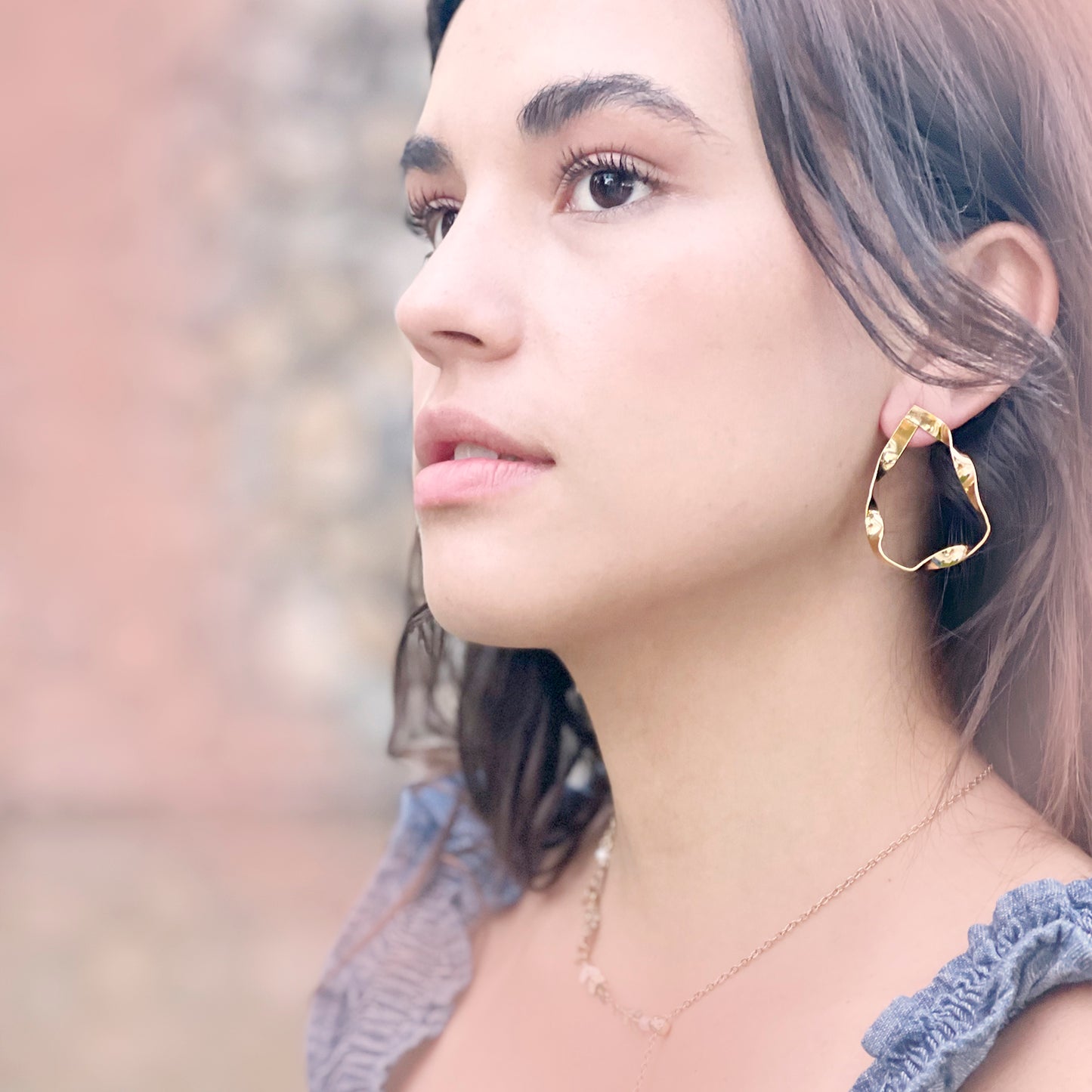 A woman with natural makeup and loose wavy hair gazes into the distance while wearing a single, large gold-tone statement earring. The earring features an abstract, wavy teardrop shape with a hammered texture that reflects light. She is dressed in a soft blue ruffled top and a delicate gold chain necklace. The background is softly blurred, highlighting the earring and her serene expression.