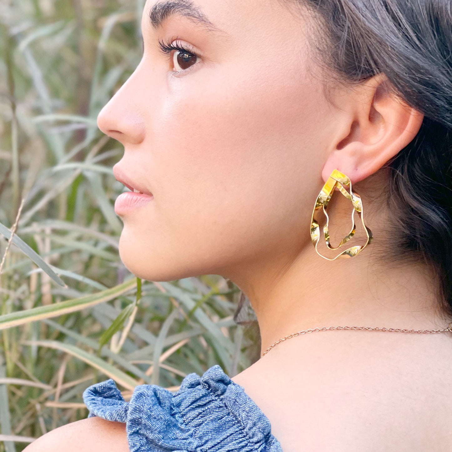 A woman with smooth, glowing skin and dark wavy hair pulled back wears a bold, sculptural gold earring shaped like a layered teardrop with fluid, organic edges. She is dressed in a blue textured top with ruffle details and a delicate gold chain necklace. The background features tall, soft-focus grass, adding a natural, serene ambiance that complements the earring's artistic, nature-inspired design.