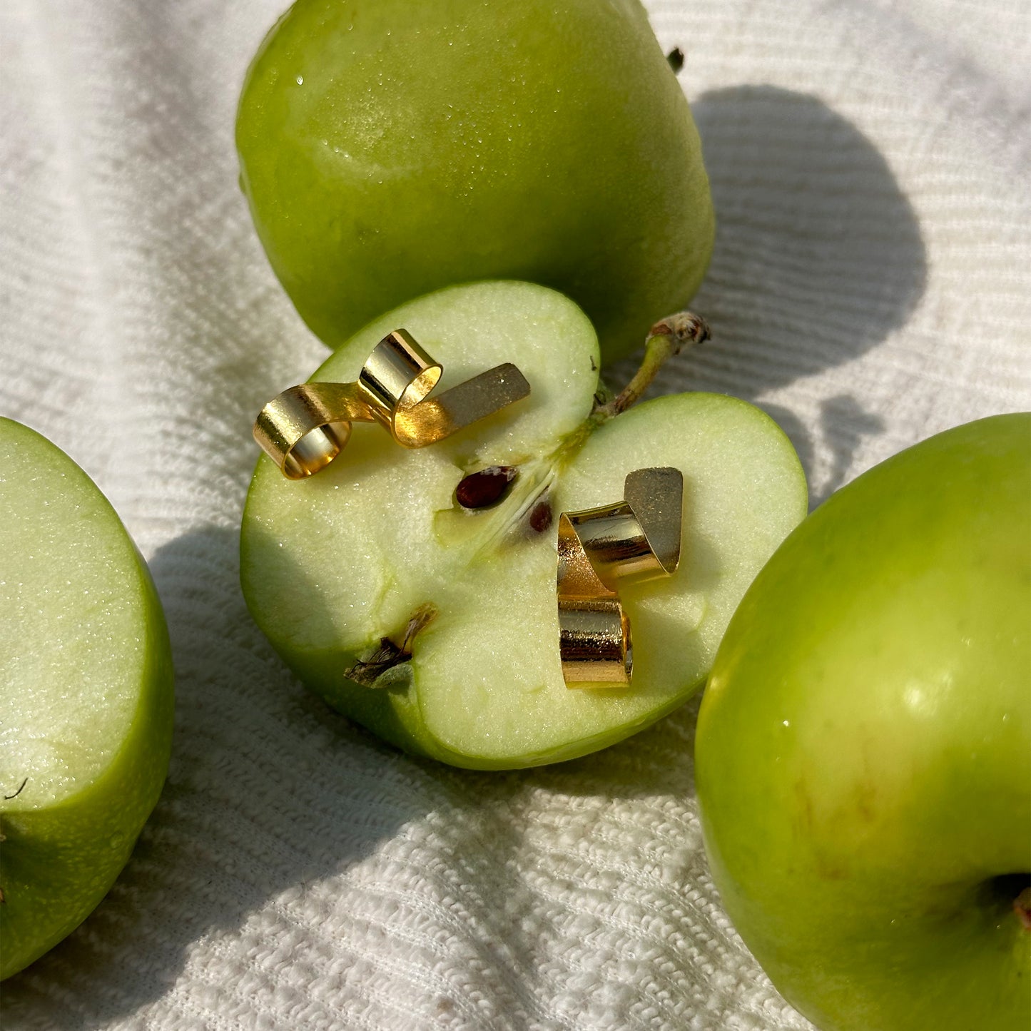 Con Cariño Earrings - Sculptural gold earrings with bold, curling ribbon-like forms are playfully styled on a fresh cut green apple. Surrounded by fresh green apples and textured neutral napkins, the composition blends modern jewelry design with fresh, minimal tabletop elements and natural light for a whimsical, editorial presentation.