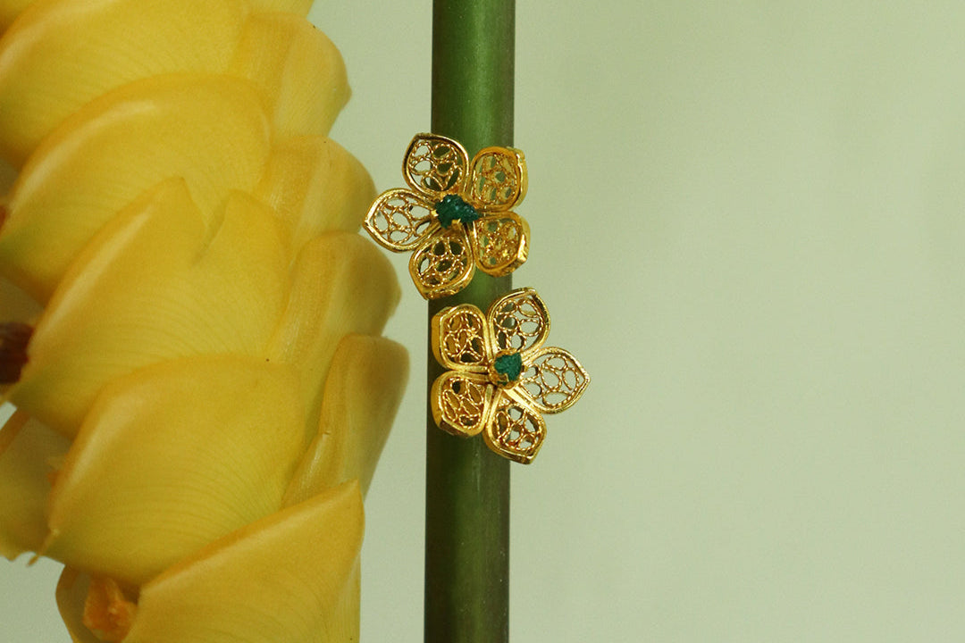 Con Cariño Earrings - Gold floral stud earrings with delicate filigree petals and green centers, displayed on a tall green stem beside a bright yellow tropical flower, highlighting intricate handcrafted detail.