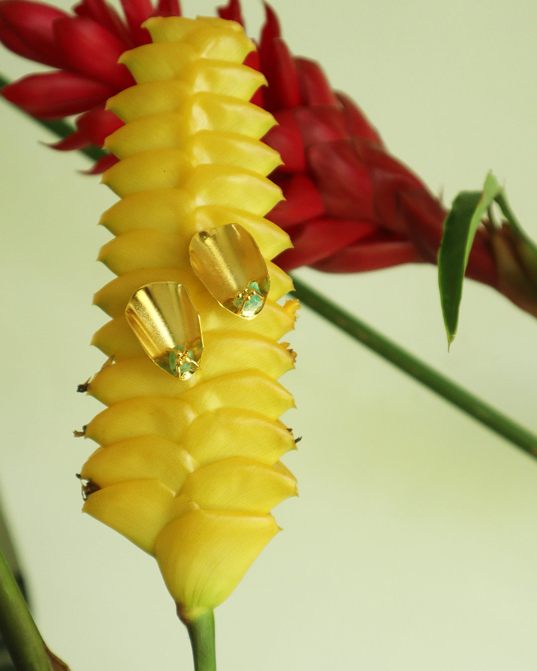 Con Cariño Earrings - Gold petal-shaped earrings with raw green stones displayed on a vibrant yellow tropical flower, with red blooms in the background, highlighting bold, botanical-inspired jewelry design.