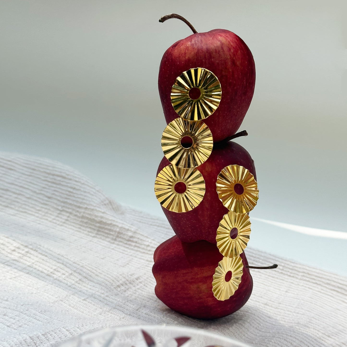 Con Cariño Earrings - Radiant gold sunburst earrings with concentric ridged discs and central cutouts artfully displayed on a vertical stack of three red apples. The setup sits on a textured white cloth with soft lighting, blending playful styling with bold, sculptural jewelry for a striking and modern composition.