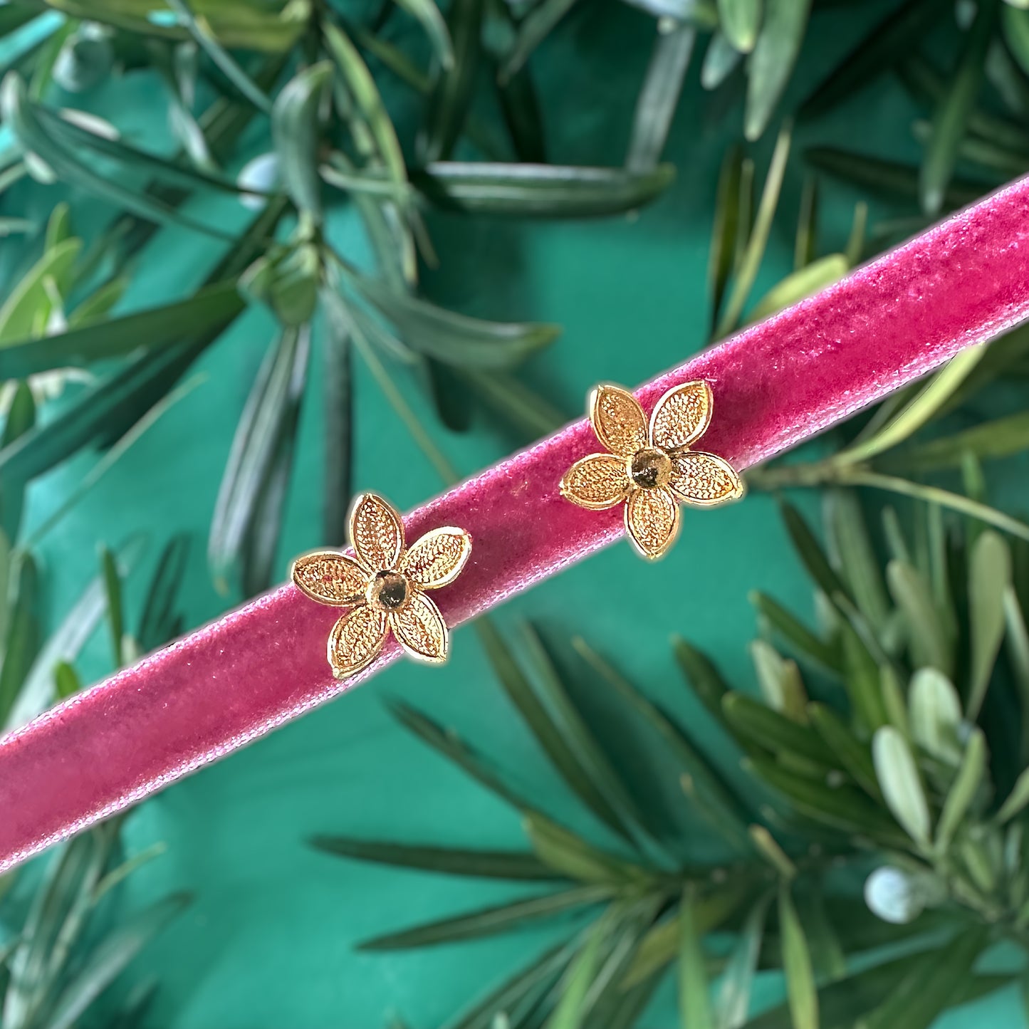 Con Cariño Earrings - Gold flower-shaped stud earrings with filigree detailing and dark center stones, displayed on a pink velvet ribbon with a green botanical background, creating a festive and elegant presentation.