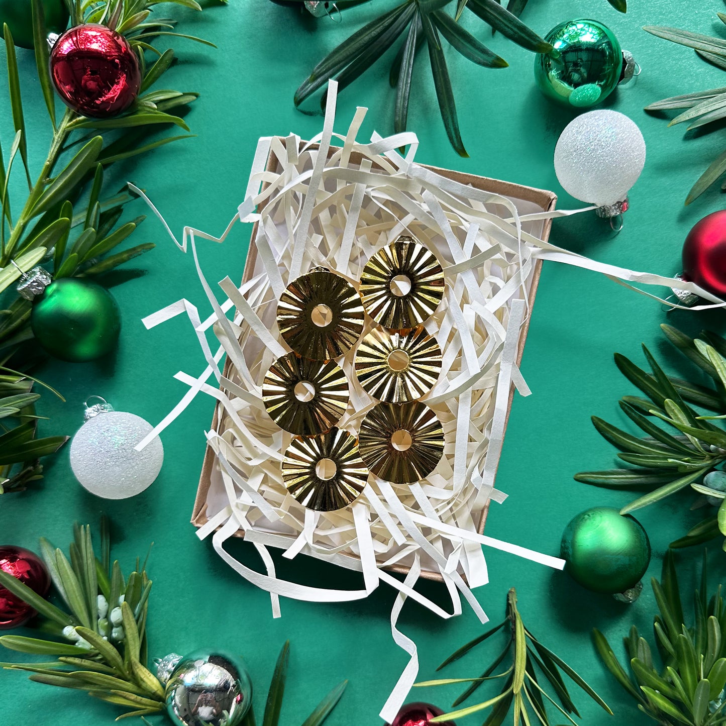 Con Cariño Earrings - Shiny gold sunburst earrings with radiating ridges and circular cutouts presented in a festive gift box filled with white crinkle paper. The box is surrounded by evergreen sprigs and colorful Christmas ornaments on a green background, creating a joyful and elegant holiday-themed display.