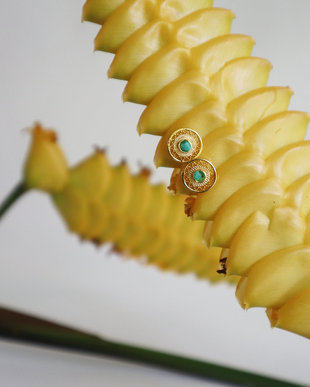 Con Cariño Earrings - Round gold stud earrings with intricate detailing and green center stones, displayed on a vibrant yellow tropical flower, highlighting handcrafted elegance and bold color contrast.