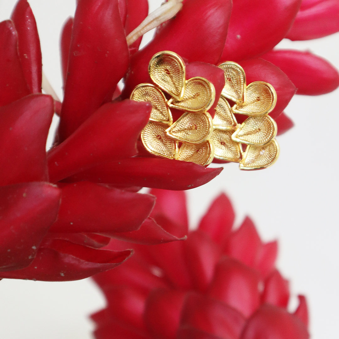 Con Cariño Gold stud earrings with layered teardrop-shaped petals arranged in a floral design, displayed on vibrant red ginger flowers for a bold and tropical aesthetic.