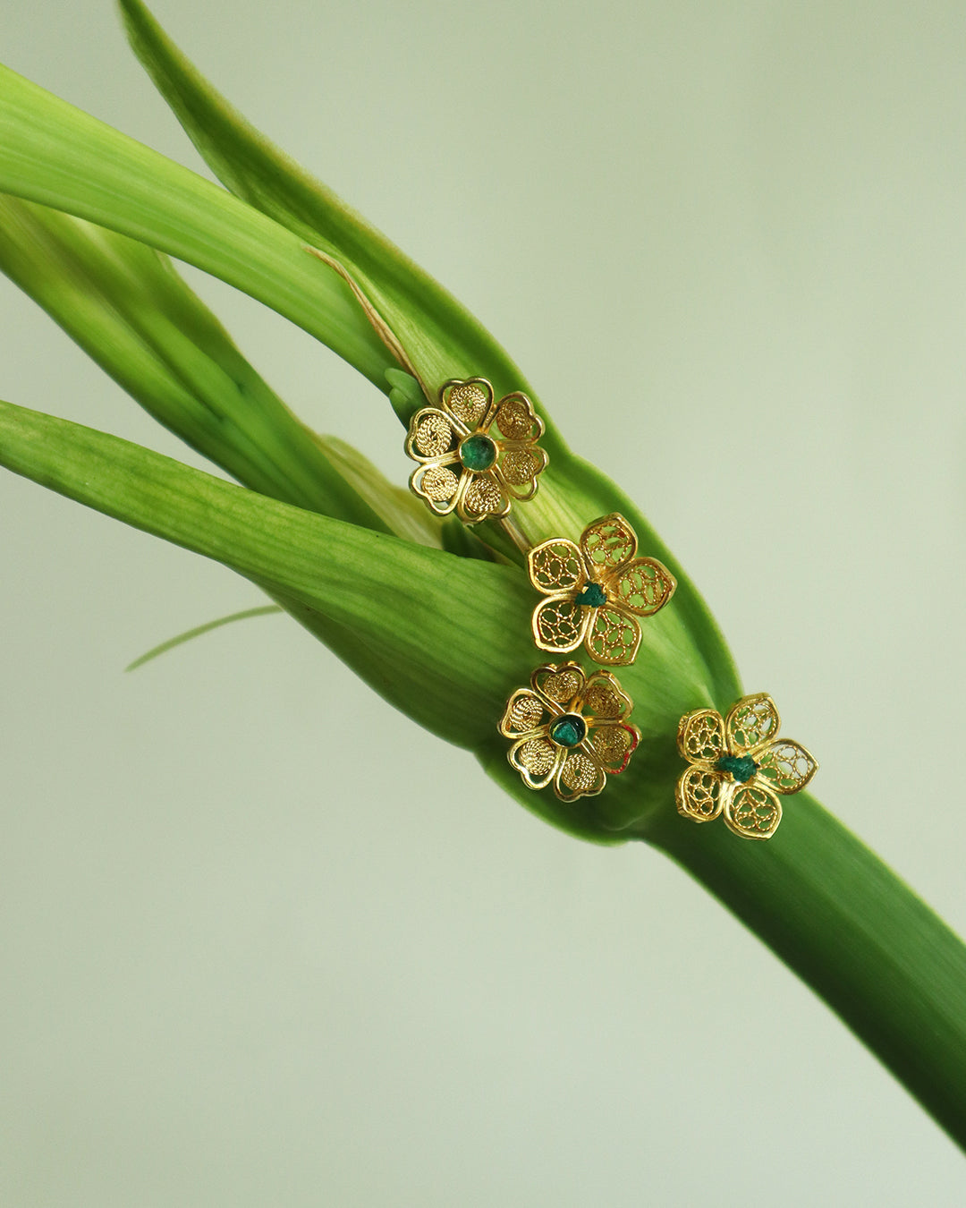 Con Cariño Earrings - Delicate gold flower-shaped stud earrings with filigree detailing and green center stones, artistically arranged on a fresh green lily stem, showcasing handmade Colombian craftsmanship.