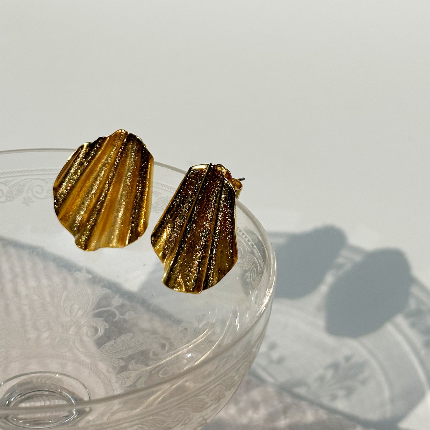 A pair of gold-toned Con Cariño earrings with sculptural, fan-like folds and a shimmering finish rest delicately on the rim of an etched vintage coupe glass. The glass casts soft floral-patterned shadows onto the white surface below, while the earrings catch natural light, emphasizing their textured metallic glow in an elegant and minimalist setting.
