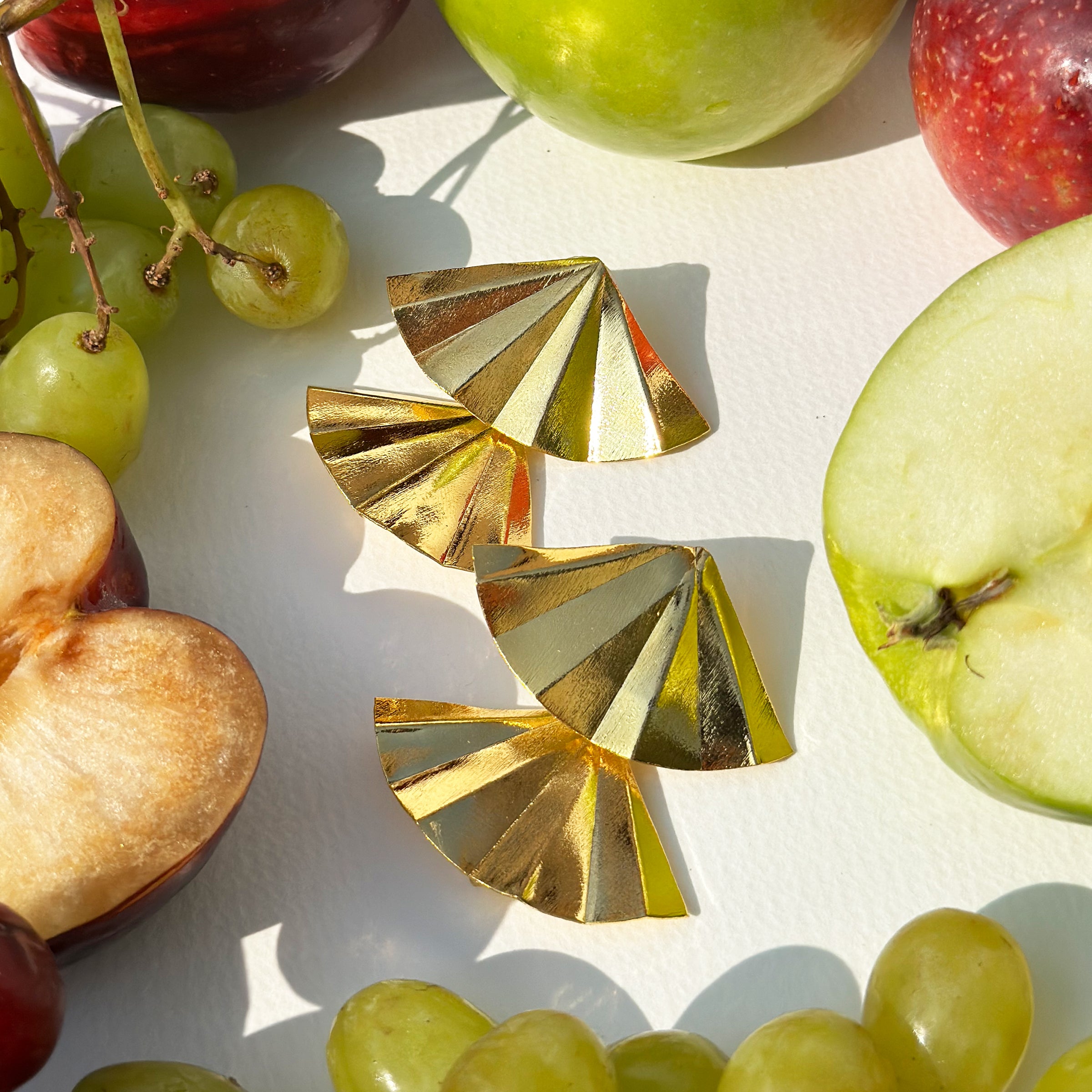 Con Cariño Earrings - Bold gold fan-shaped earrings with a polished, sculptural design displayed flat on a white surface, surrounded by an assortment of fresh fruit including sliced apples, grapes, and plums. The vibrant colors and natural textures of the fruit contrast beautifully with the sleek metallic shine of the earrings, creating a playful yet luxurious composition.