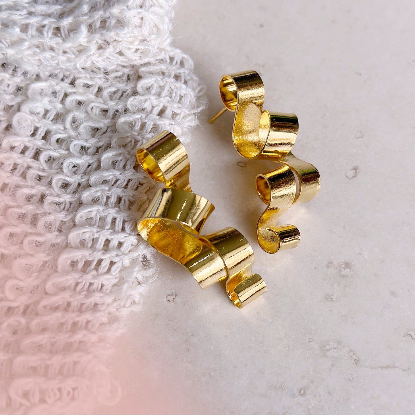 A pair of shiny gold earrings with a flowing, ribbon-like design rests elegantly on a smooth beige stone surface. One of the earrings leans against a textured, white woven fabric, creating a contrast between the soft textile and the structured metallic form. The overall setting feels warm and sophisticated, with a soft pink hue subtly blending into the corner of the composition.