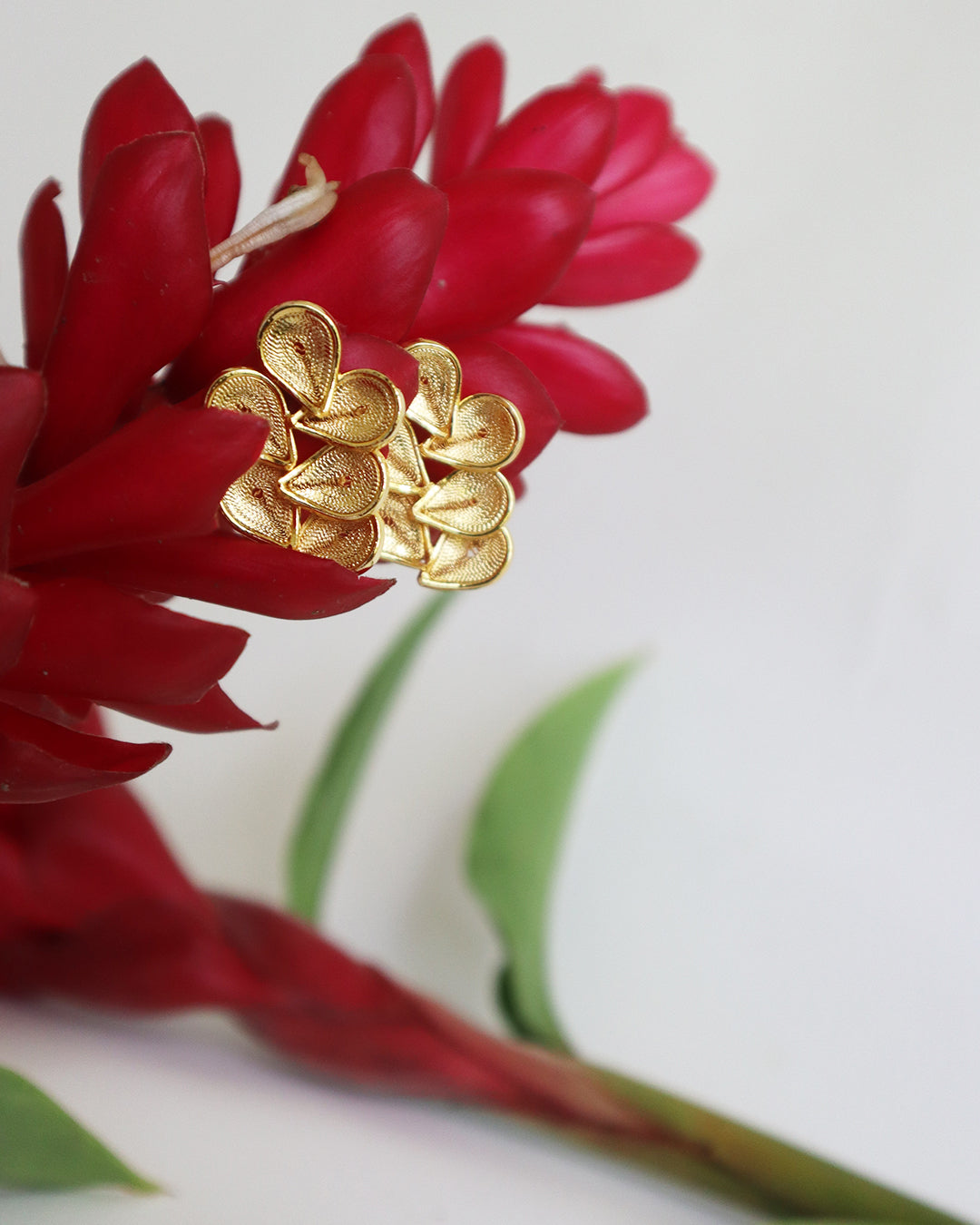Con Cariño Gold stud earrings with layered teardrop-shaped petals arranged in a floral design, displayed on vibrant red ginger flowers for a bold and tropical aesthetic.