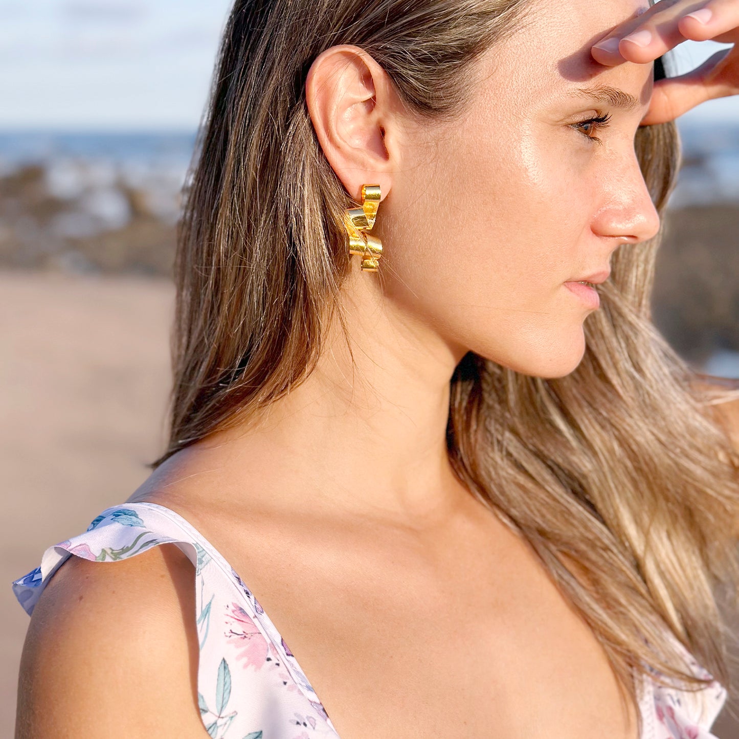 A woman with light brown hair gazes toward the sun on a beach, shielding her eyes with one hand. She wears sculptural gold earrings that resemble curled ribbons. Her floral dress features delicate pastel tones, and the soft golden hour lighting accentuates her skin's natural glow, adding a serene and elegant feel to the coastal setting.