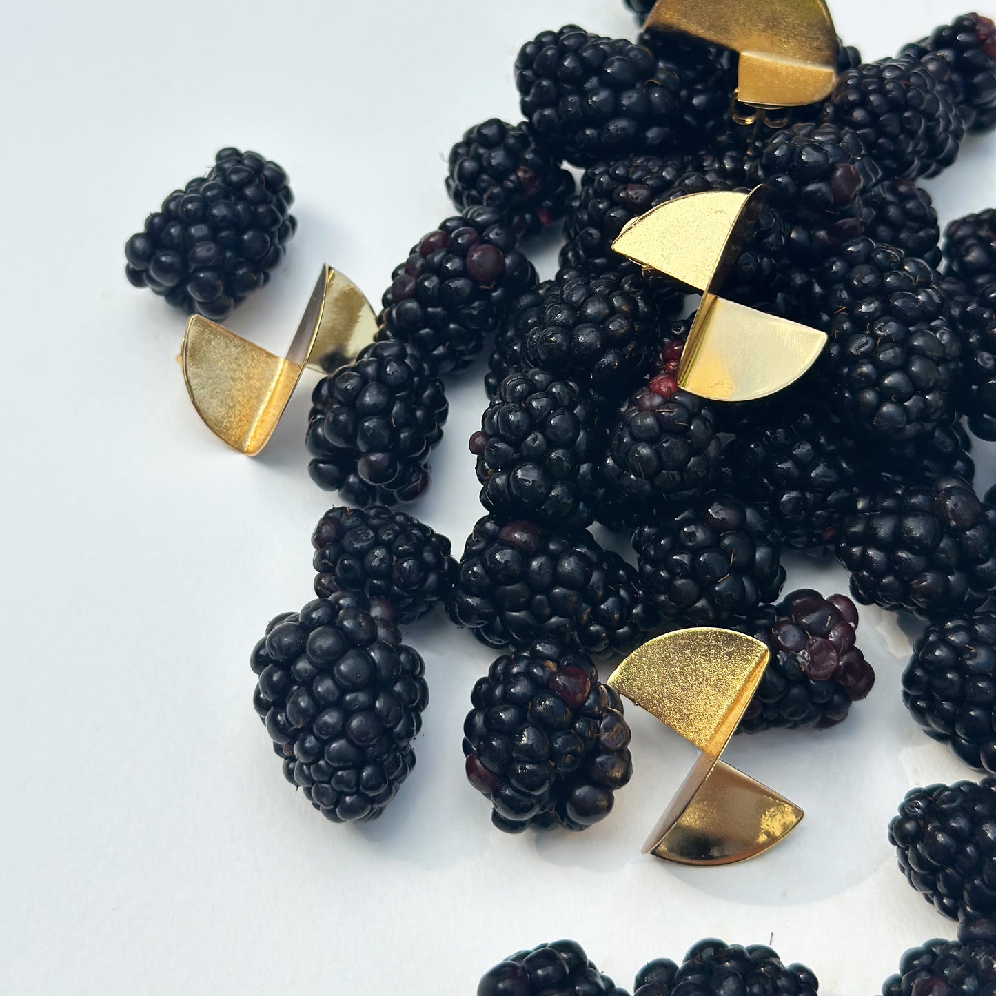 A group of geometric gold stud earrings, each formed by two joined semi-circles, is artfully scattered among fresh, ripe blackberries on a clean white surface. The polished gold surfaces contrast vividly with the deep purple tones and textured skin of the berries, creating a bold yet elegant visual composition that blends luxury with natural elements.