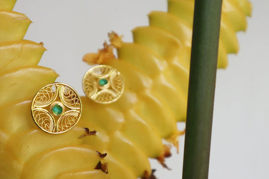 Intricate gold filigree stud earrings with green center stones styled on yellow tropical blooms.