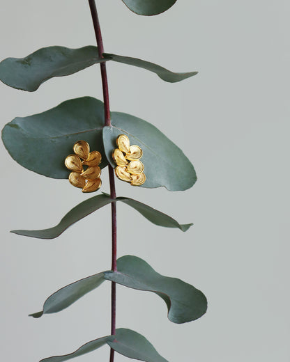 Elegant gold earrings featuring five overlapping teardrop-shaped petals with fine engraved texture, displayed on muted green eucalyptus leaves against a soft gray background, creating a refined and nature-inspired presentation that highlights the handcrafted details and warm metallic finish.
