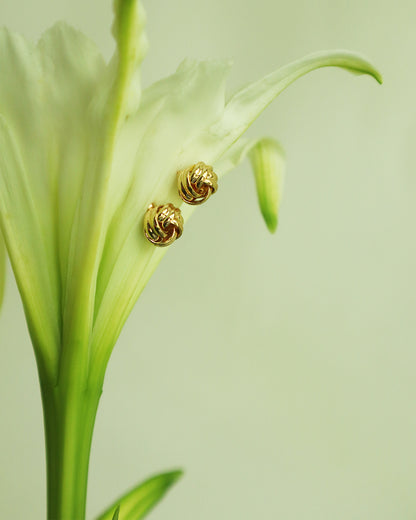 Con Cariño Earrings - Gold knot stud earrings gently placed on the petal of a white lily, highlighting their elegant woven design against a soft botanical background.
