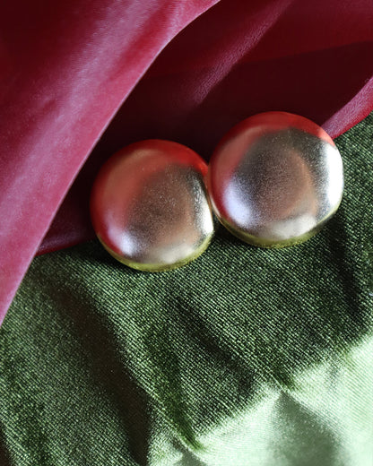 Round gold stud earrings with a smooth, domed finish, displayed on layered green and deep red satin fabric.