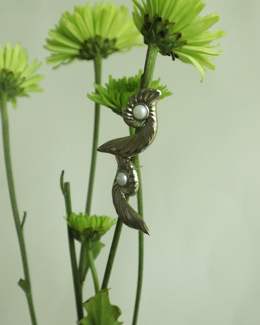 Silver-toned sculptural earrings with pearl accents and flowing, wing-like curves displayed on tall green stems, blending modern elegance with organic inspiration.