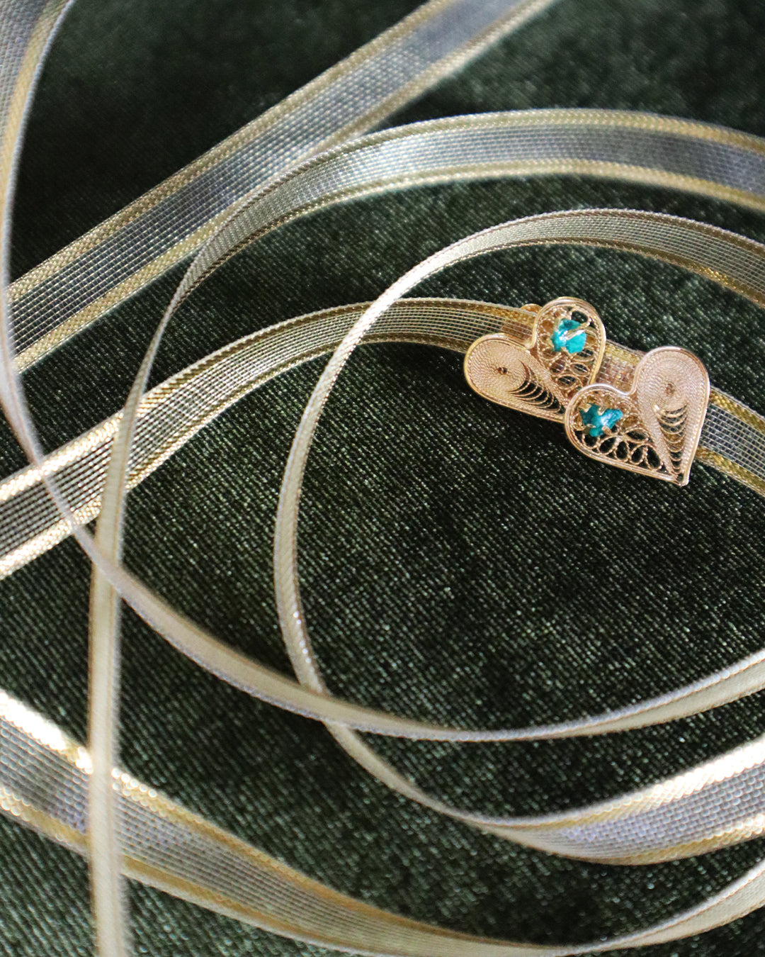Delicate gold filigree heart-shaped earrings with small turquoise stones in the center, displayed on dark green textured fabric and intertwined with sheer gold ribbon loops.