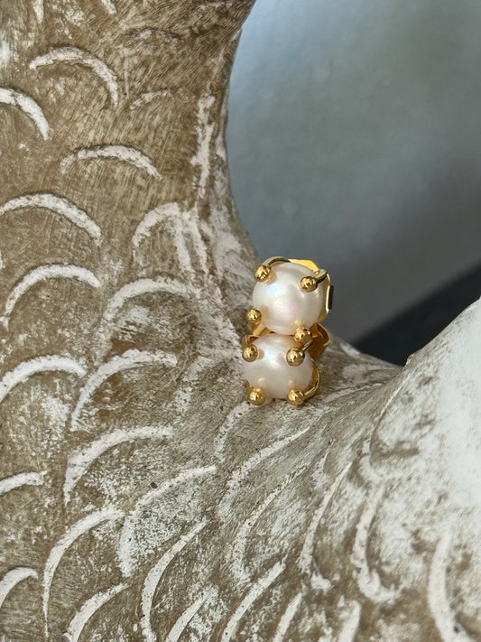 Gold stud earring featuring two stacked white pearls secured with rounded gold prongs, displayed against a textured beige sculpture with carved wave-like patterns.