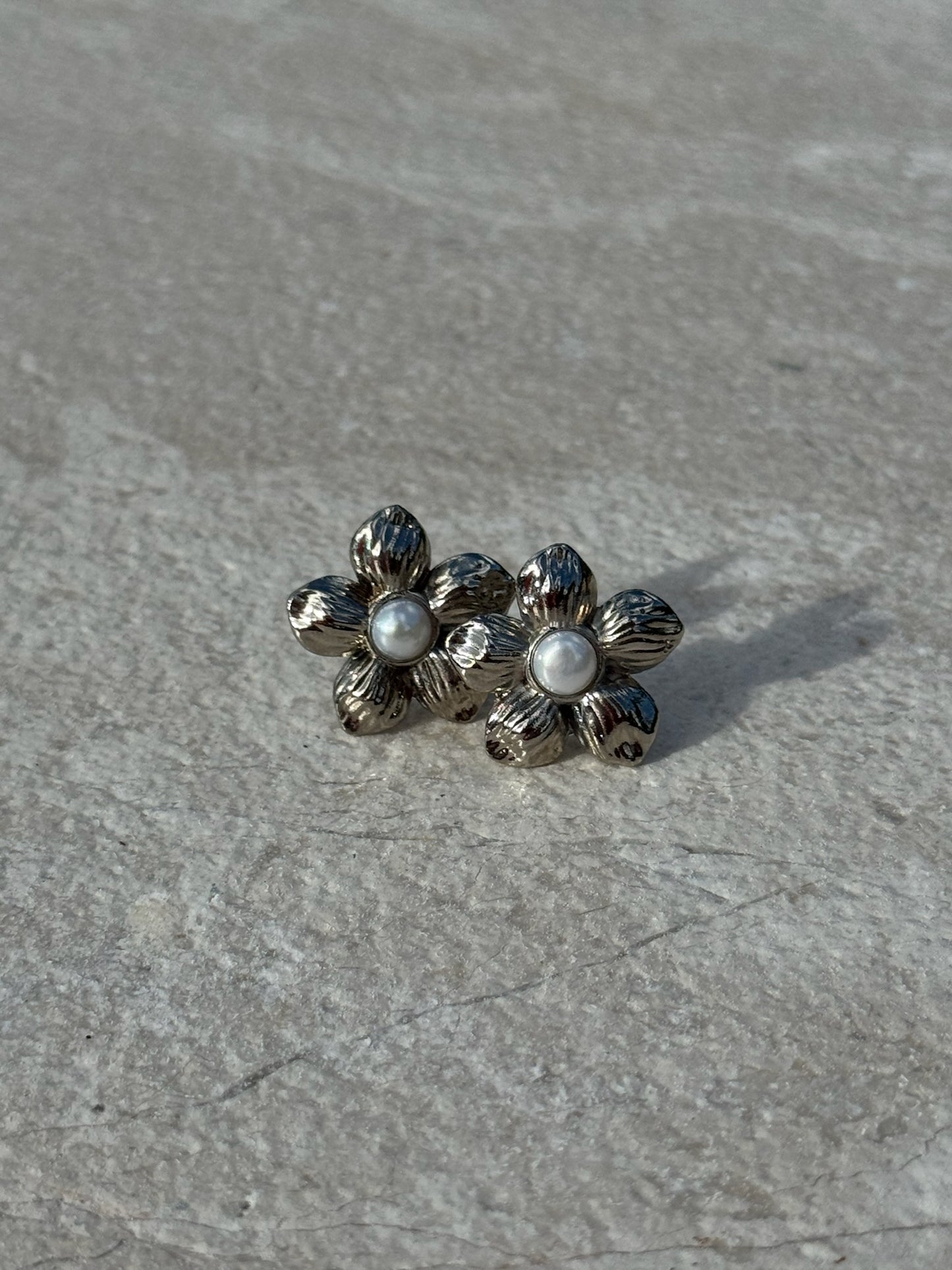 Silver-toned flower-shaped stud earrings with textured petals and a small white pearl at the center of each, displayed on a light stone surface.