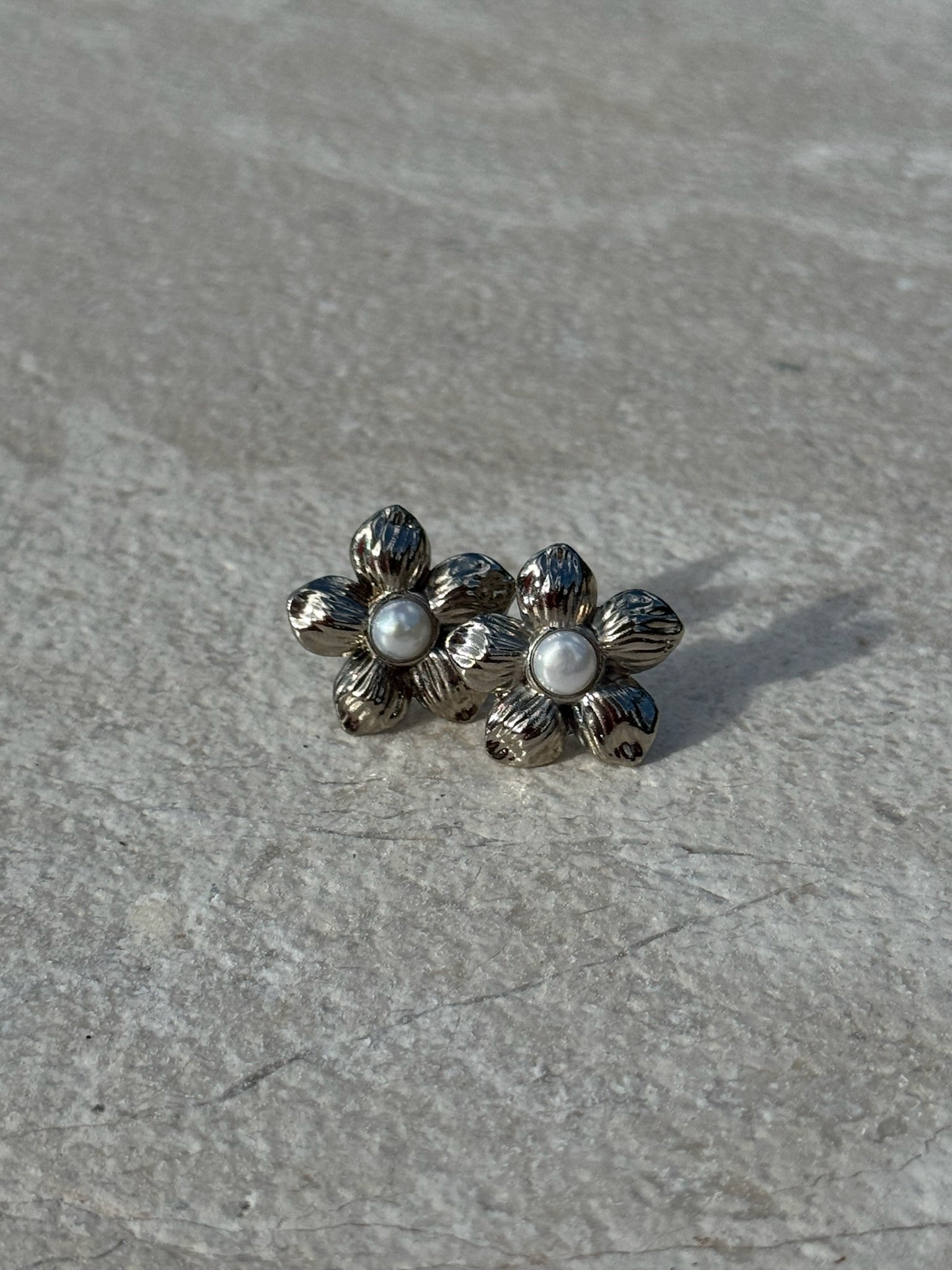 Silver-toned flower-shaped stud earrings with textured petals and a small white pearl at the center of each, displayed on a light stone surface.