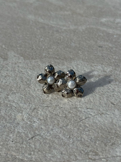 Silver-toned flower-shaped stud earrings with textured petals and a small white pearl at the center of each, displayed on a light stone surface.