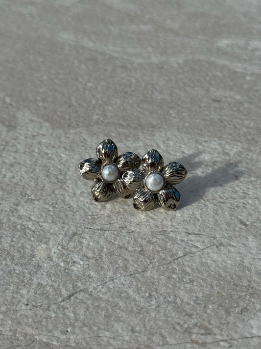Silver-toned flower-shaped stud earrings with textured petals and a small white pearl at the center of each, displayed on a light stone surface.