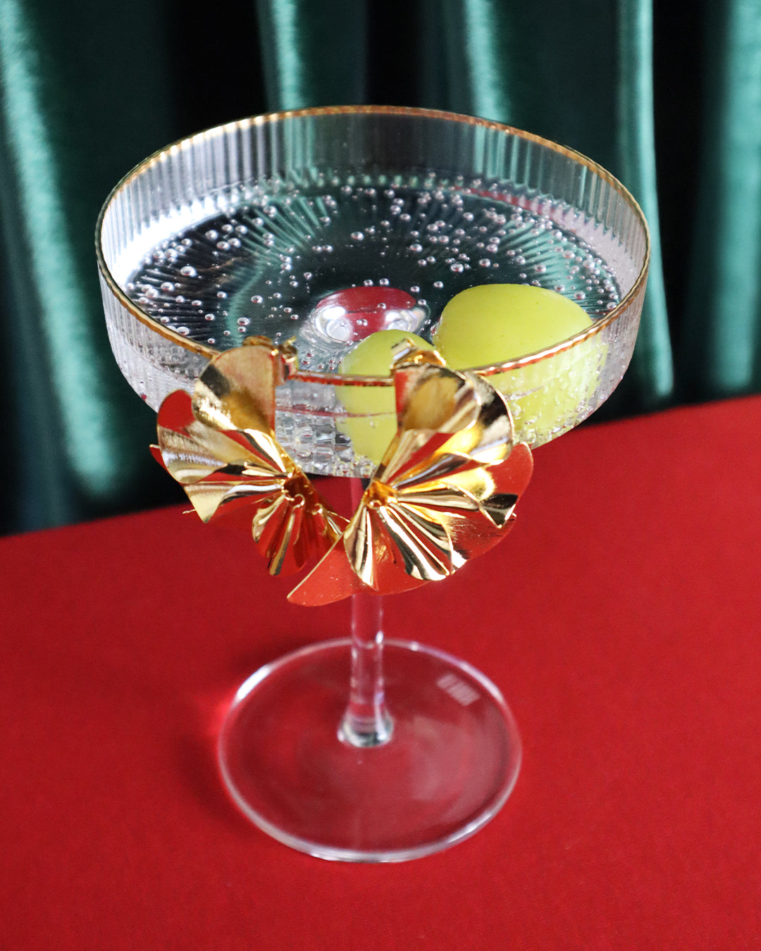 Large gold statement earrings with sculptural, fan-like folds hanging over the rim of a glass coupe filled with a sparkling drink and green grapes, set against a deep green backdrop and red surface.