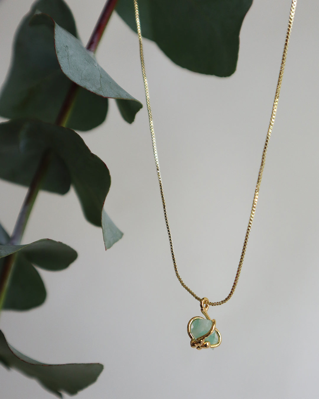  Raw emerald organic pendant on a fine gold chain, hanging gracefully in front of eucalyptus leaves.