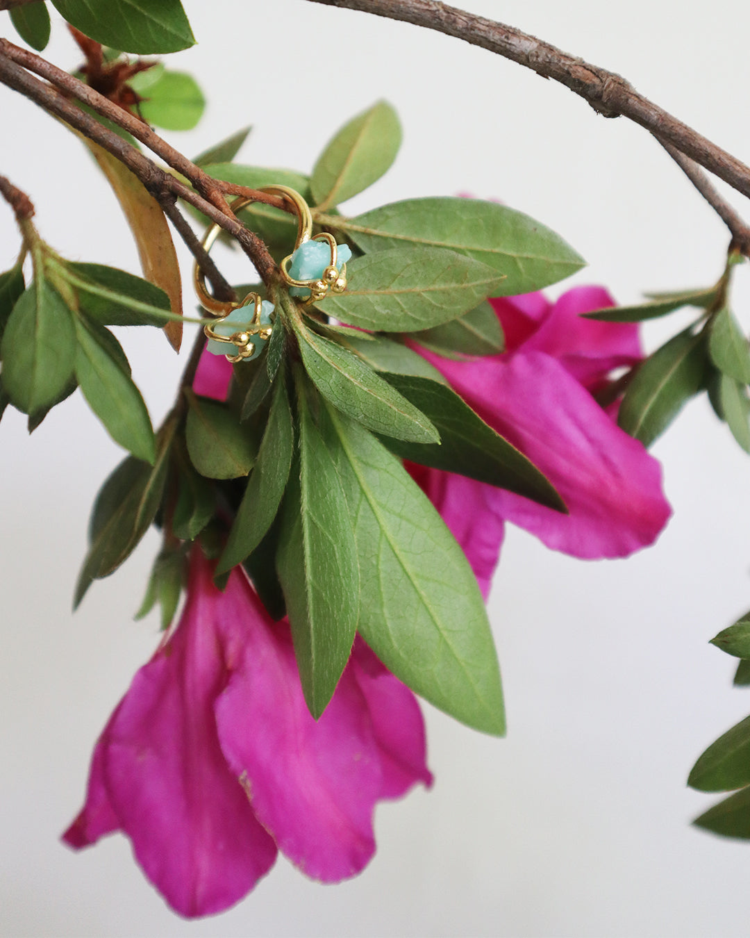 24K gold-dipped ring with Raw emerald stones nestled in fresh green leaves and vibrant magenta flowers.
