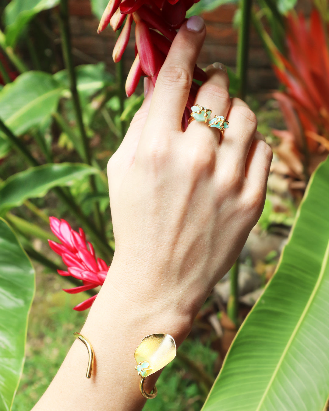 Hand adorned with a gold bracelet featuring a petal-shaped design and raw Emerald stone, paired with a matching gold ring with turquoise stones, set against a tropical background of red ginger flowers and lush green leaves.