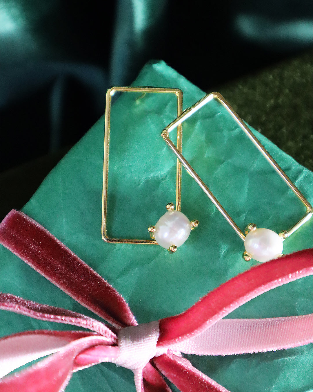 Gold rectangular drop earrings featuring a single small white pearl at the base of each frame, displayed on teal wrapping paper tied with a velvet pink ribbon.