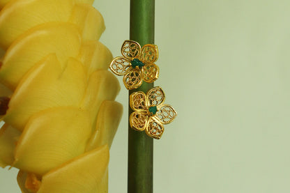 Con Cariño Earrings - Gold floral stud earrings with delicate filigree petals and green centers, displayed on a tall green stem beside a bright yellow tropical flower, highlighting intricate handcrafted detail.