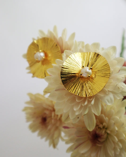 Con Cariño Earrings- Bold gold disc earrings with a radiant textured finish and central pearl accent, beautifully displayed on soft cream-colored chrysanthemum flowers.