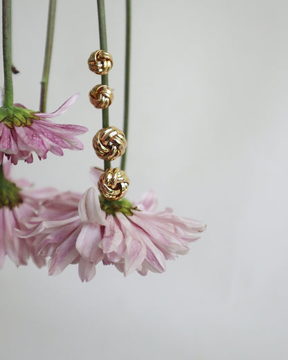Con Cariño Earrings - Gold knot stud earrings in various sizes styled on soft pink chrysanthemums, highlighting their elegant texture and timeless design against a neutral background.