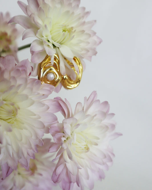 Con Cariño Earrings -  Sculptural gold earrings with an abstract design and pearl center, delicately placed among soft pink and white chrysanthemum flowers, highlighting their elegant and artistic form.